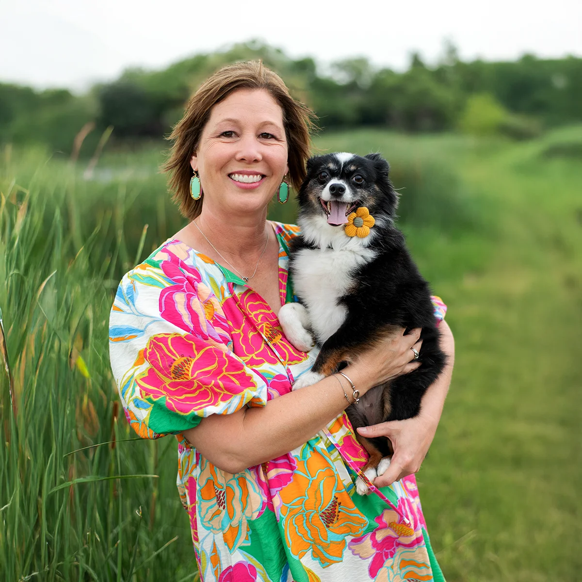 Woman smiling and holding a small black and white dog wearing a flower accessory, standing in a grassy field with tall reeds and trees in the background.